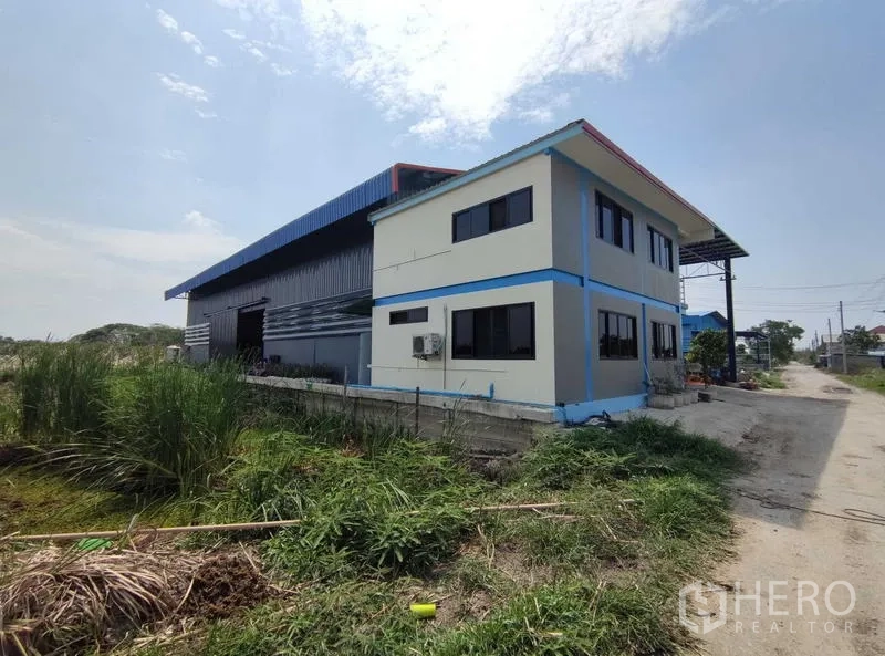 Warehouse for sale in Nakhon Pathom - Rear angle of the metal‑clad warehouse beside a two‑storey office block with surrounding greenery and service lane.