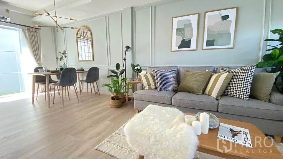 Townhouse for rent in Prawet, Bangkok - Cozy lounge with gray sofa, patterned cushions and coffee table next to a dining set under a gold chandelier.