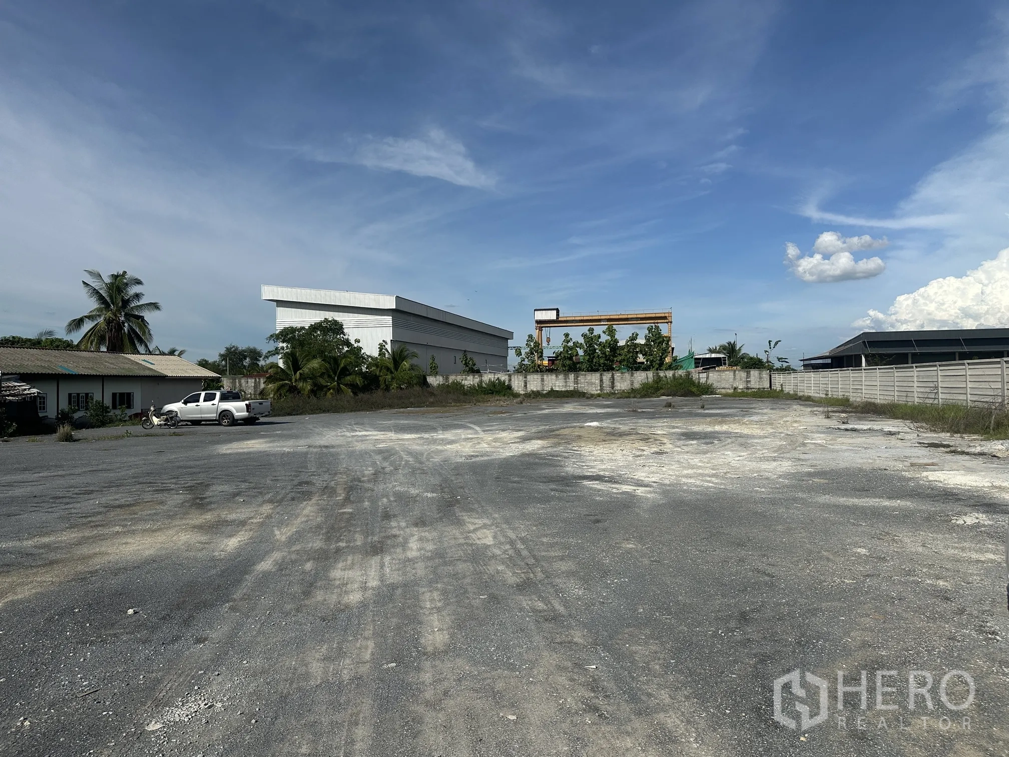 Land for sale in Sai Noi, Nonthaburi - Level gravelled land plot with boundary wall, nearby warehouse and a parked pickup truck.