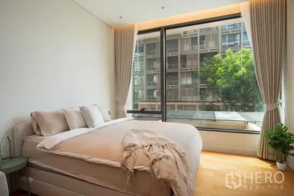 คอนโด ให้เช่าในPathum Wan, Bangkok - ห้องนอนริมหน้าต่างบานใหญ่ ชุดเครื่องนอนโทนอุ่น ผ้าม่านเต็มความสูง มองเห็นอาคารเมืองและต้นไม้