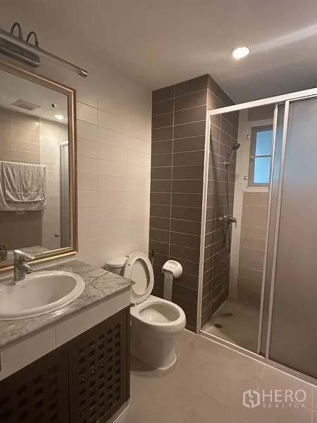 Condominium for sale in Watthana, Bangkok - Modern bathroom with marble-top vanity, toilet and separate glass-enclosed shower with brown accent tiles.