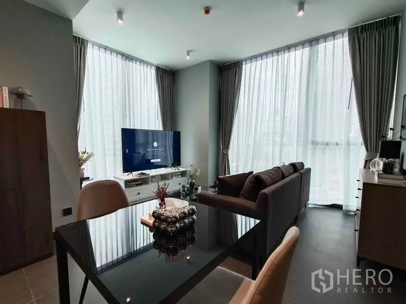 Condominium for rent in Bang Rak, Bangkok - Open-plan living and dining area with floor-to-ceiling windows, grey sofa, TV, and a glass dining table at Tait Sathorn 12.
