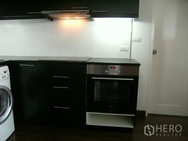 Condominium for rent in Watthana, Bangkok - Modern kitchen detail showing built-in oven, cooktop, hood, and a washing machine.