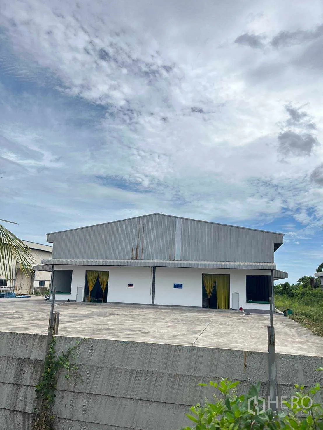 工厂 出售在Nikhom Phatthana, Rayong - 仓库立面，遮棚装卸区与围墙后抬高的混凝土场地。