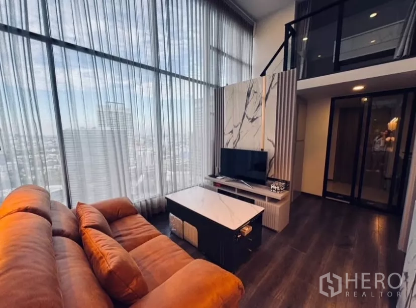 Condominium for rent in Chatuchak, Bangkok - Double-height living room with city view, brown sofa, TV, and mezzanine in a duplex condo.