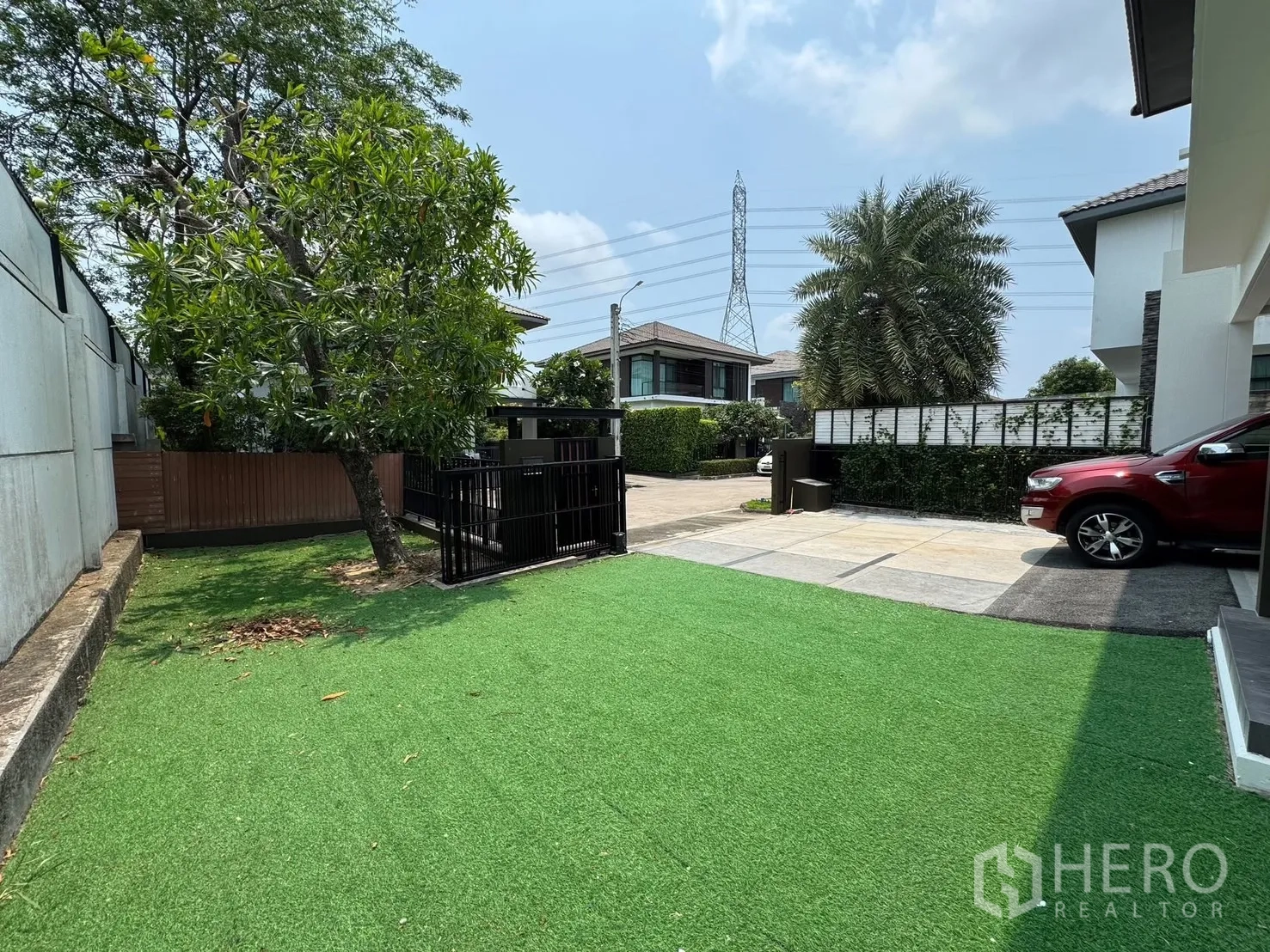 Detached House for rent in Prawet, Bangkok - Front garden with artificial turf, a mature tree and view toward the entrance gate.