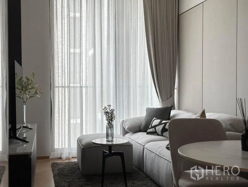 Condominium for rent in Watthana, Bangkok - Bright living room with floor-to-ceiling sheer curtains, a light gray sectional sofa, round side table and TV console.