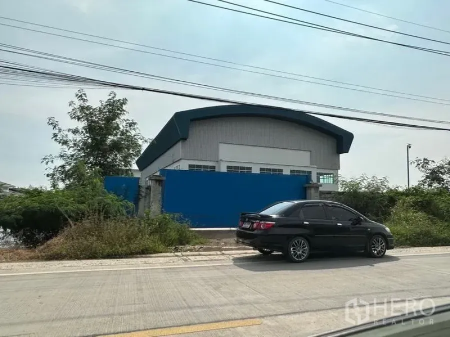 工厂 出租在Pak Kret, Nonthaburi - 临街工厂外观，蓝色围墙与弧形金属屋顶，靠混凝土道路。