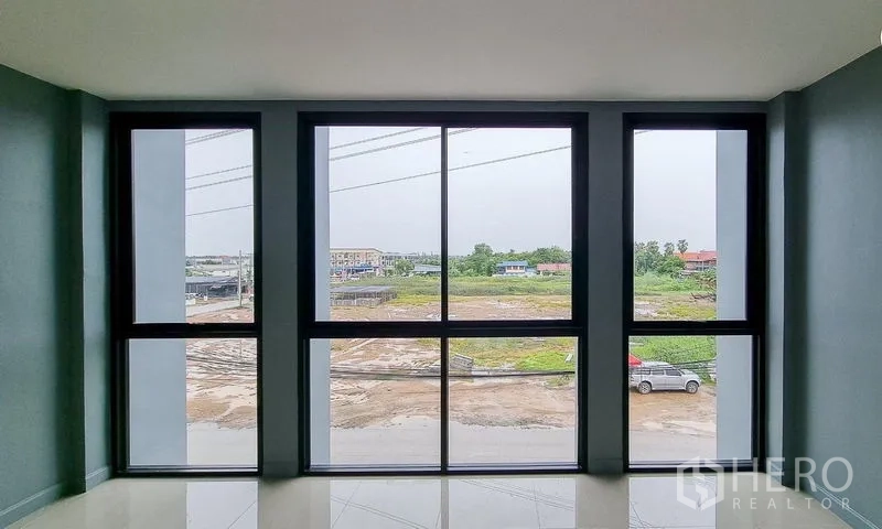 Shophouse for sale in Lam Luk Ka, Pathum Thani - View through tall black-framed windows to the road and open land outside.