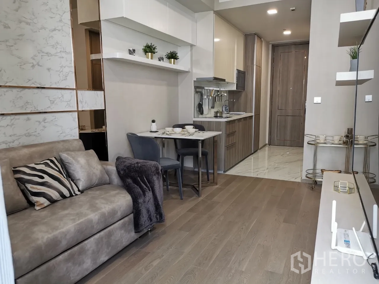Condominium for rent in Watthana, Bangkok - Modern open-plan living room with sofa, dining table, kitchenette, wood floors, and floating shelves.