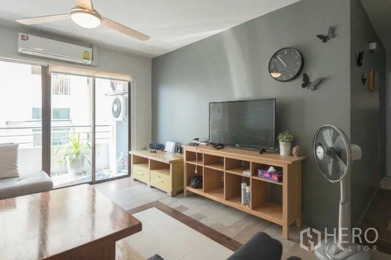 คอนโด ให้เช่าในWatthana, Bangkok - ห้องนั่งเล่นพร้อมชั้นวางทีวี ผนังเทา พัดลมเพดาน และประตูกระจกบานเลื่อนเปิดสู่ระเบียง