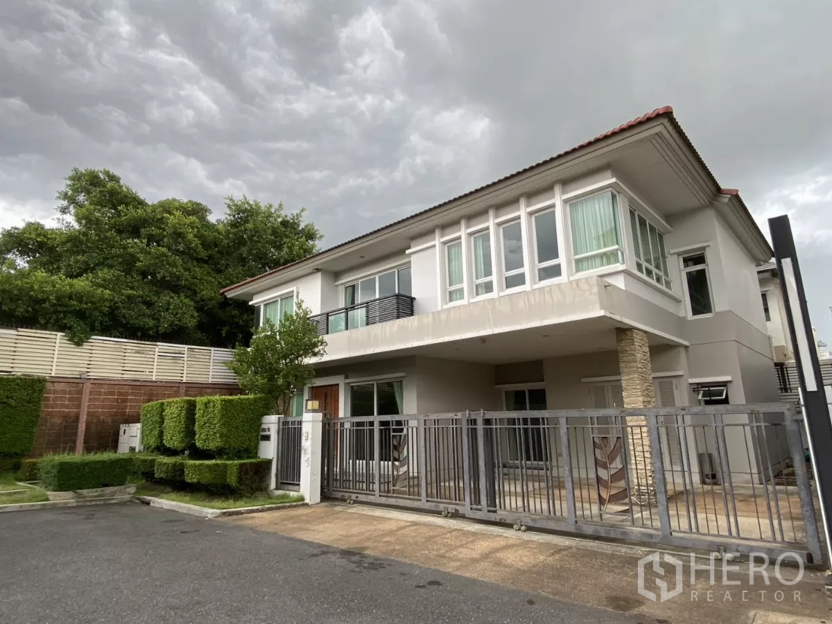 บ้าน ให้เช่าหรือขายในLak Si, Bangkok - บ้านเดี่ยวสองชั้นสไตล์โมเดิร์น พร้อมรั้วหน้าบ้านและพุ่มไม้ตัดแต่ง ท่ามกลางท้องฟ้ามีเมฆ.