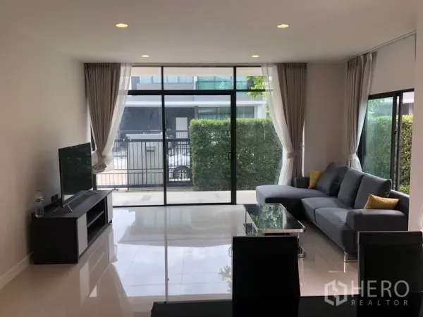 Detached House for rent or sale in Bang Kapi, Bangkok - Bright living room with floor-to-ceiling windows, gray sectional sofa and TV console.