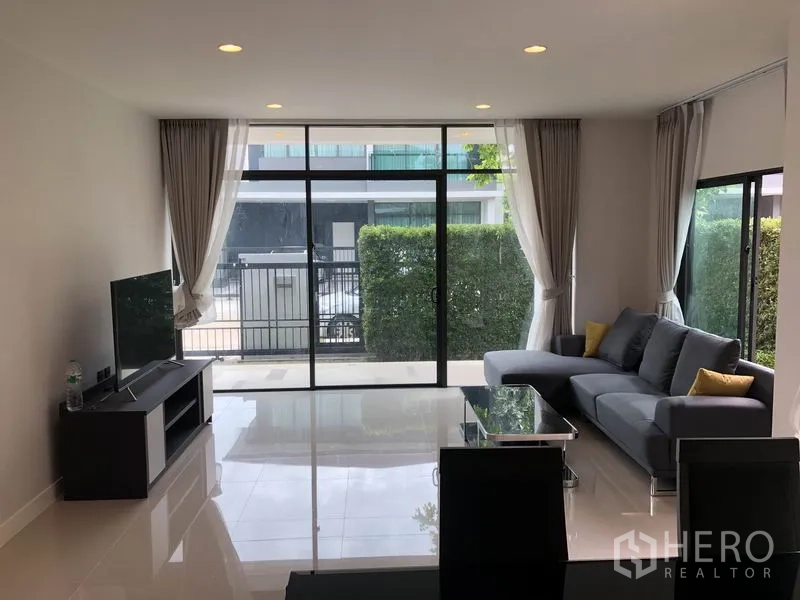 Detached House for rent or sale in Bang Kapi, Bangkok - Bright living room with floor-to-ceiling windows, gray sectional sofa and TV console.