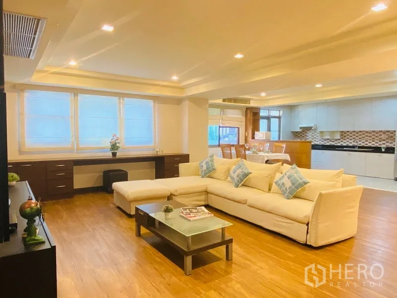 Condominium for rent in Watthana, Bangkok - Spacious open-plan living area with a white L-shaped sofa, glass coffee table, wood floors and a bright kitchen with mosaic backsplash.