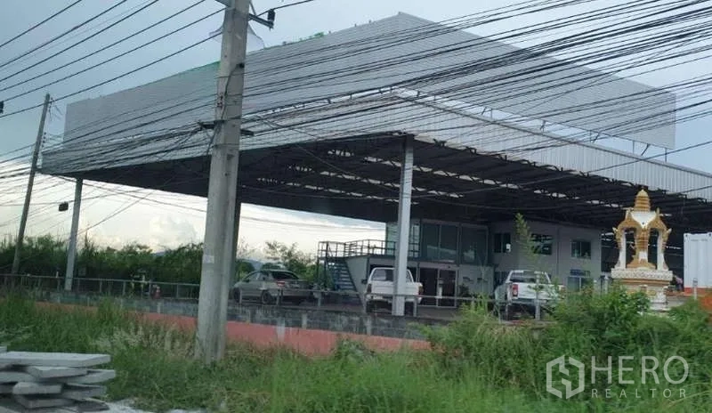 Land for sale in Si Racha, Chon Buri - Steel-frame covered yard and office building viewed from the roadside, with parked vehicles, greenery, and a small shrine.