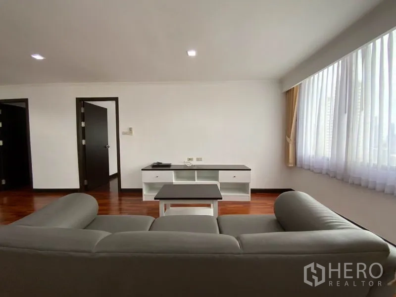 คอนโด ให้เช่าในWatthana, Bangkok - ห้องนั่งเล่น โซฟาสีเทาหันหน้าไปยังชั้นวางทีวี ใต้หน้าต่างบานกว้าง พื้นไม้ขัดเงา