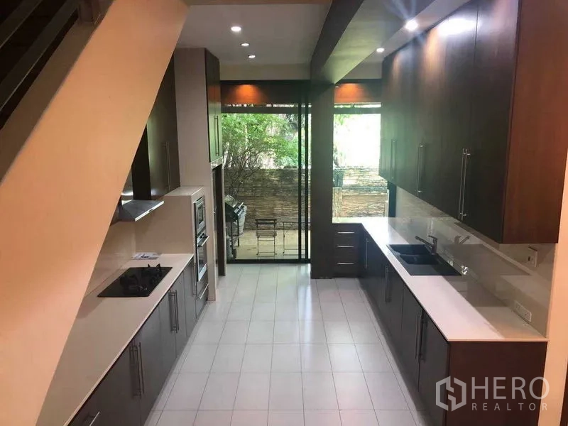 Townhouse for rent in Khlong Toei, Bangkok - Long kitchen with twin counters, built‑in appliances, and sliding glass doors opening to a patio.