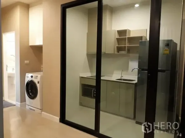 Condominium for rent in Huai Khwang, Bangkok - Enclosed kitchen behind sliding glass with built-in cabinets, sink, microwave, fridge, and a corridor washing machine.