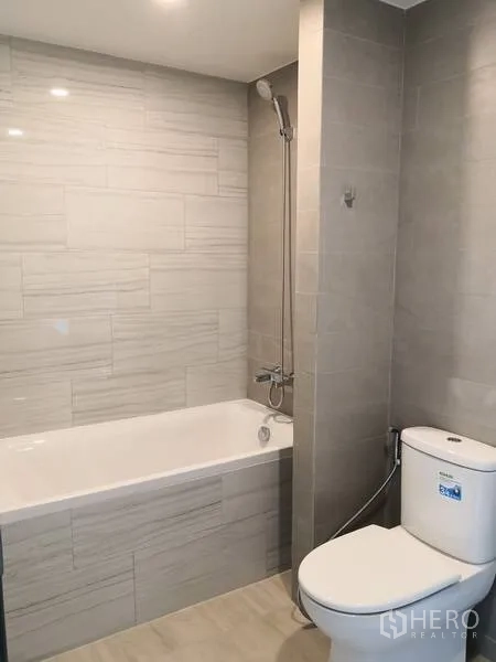 Condominium for rent or sale in Watthana, Bangkok - Modern bathroom with bathtub, shower over tub, light gray tiles, and toilet.