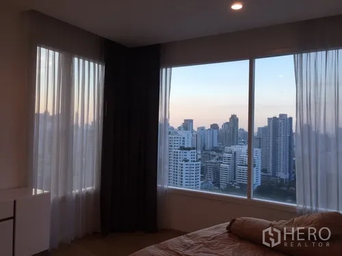 Condominium for rent in Watthana, Bangkok - Corner bedroom with large windows and panoramic Bangkok city view at sunset.