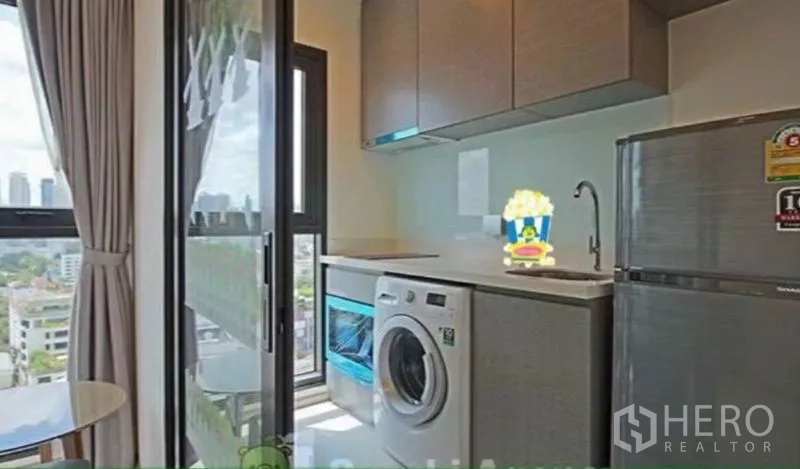 Condominium for rent in Khlong Toei, Bangkok - Compact kitchenette with washing machine, fridge and balcony door opening to Bangkok skyline views.