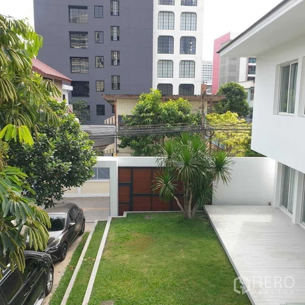 Detached House for rent in Watthana, Bangkok - Front yard with green lawn, trees and gated driveway beside a modern white house in an urban neighborhood.