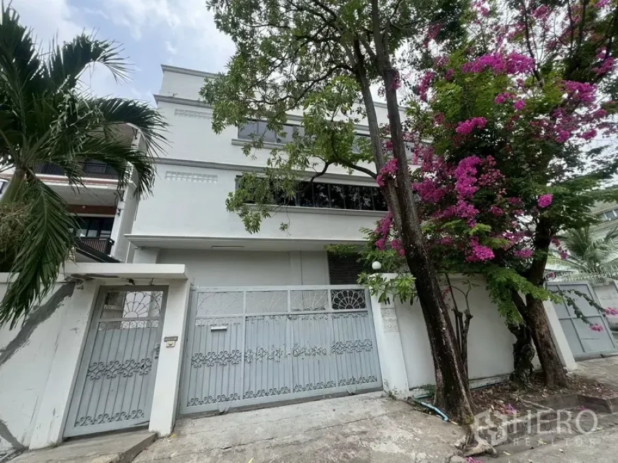 คลังสินค้า ให้เช่าในPhra Khanong, Bangkok - ด้านหน้าตัวอาคารโกดังสีขาวสามชั้น พร้อมรั้วเหล็กและต้นไม้ดอกสีชมพูในพระโขนง