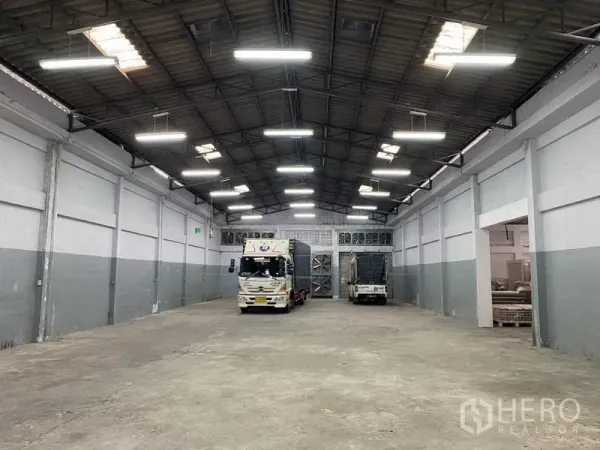 Warehouse for rent in Bang Phli, Samut Prakan - Spacious warehouse interior with high steel truss ceiling, bright LED lights and trucks at rear loading doors.