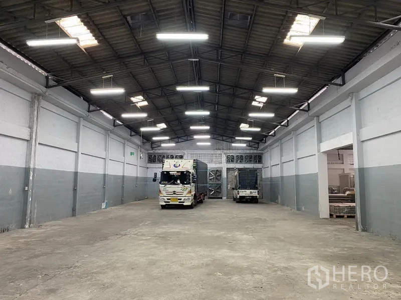 Warehouse for rent in Bang Phli, Samut Prakan - Spacious warehouse interior with high steel truss ceiling, bright LED lights and trucks at rear loading doors.