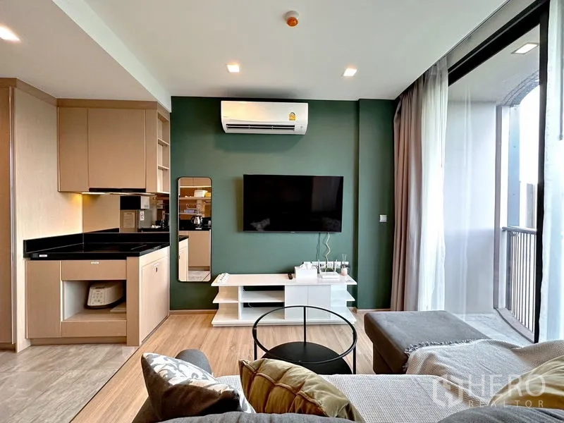 Condominium for sale in Watthana, Bangkok - Living area with green accent wall, wall‑mounted TV, and balcony access beside full‑height drapes.