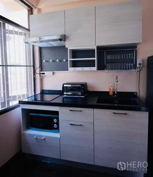 Detached House for rent in Bang Na, Bangkok - Compact light-wood kitchenette with hob, sink, range hood and appliances beside a window.