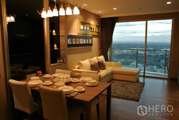 Condominium for rent or sale in Watthana, Bangkok - Warm living and dining room with L-shaped sofa, chandelier lighting, wood floors, and floor-to-ceiling glass opening to a balcony with a panoramic Bangkok skyline view.