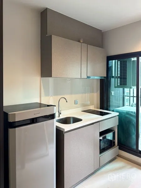 Condominium for sale in Ratchathewi, Bangkok - Compact kitchenette with fridge, sink, upper cabinets, cooktop, and microwave next to a glass door.