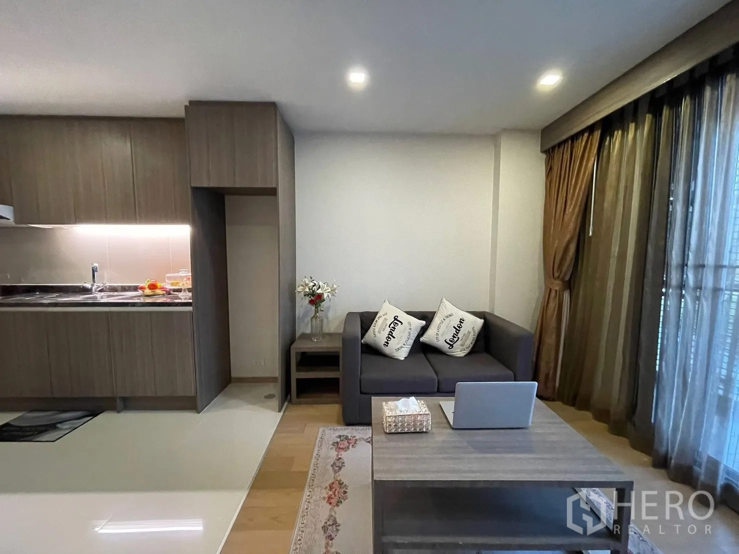 Condominium for rent in Watthana, Bangkok - Open-plan living area with gray sofa, coffee table, and a modern kitchenette with wood cabinets and under-cabinet lighting.