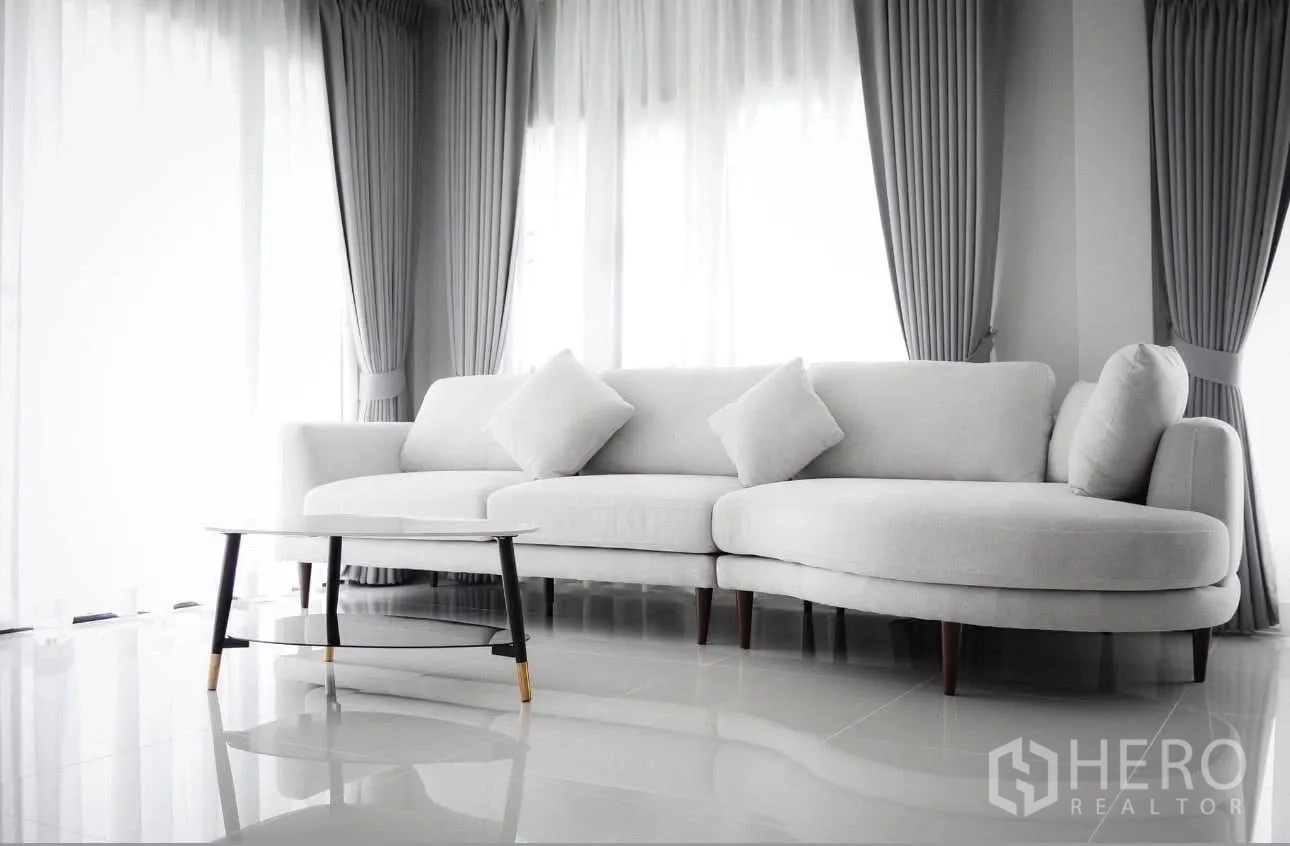 Detached House for rent in Bang Phli, Samut Prakan - White L-shaped sofa by large windows with grey curtains and a coffee table.