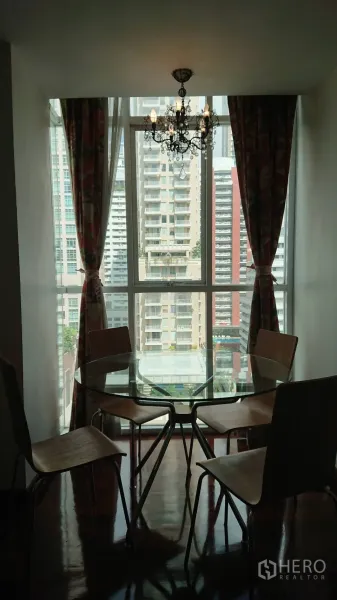 Condominium for rent in Pathum Wan, Bangkok - Dining nook with a round glass table and four chairs under a chandelier beside a large window overlooking high-rises.