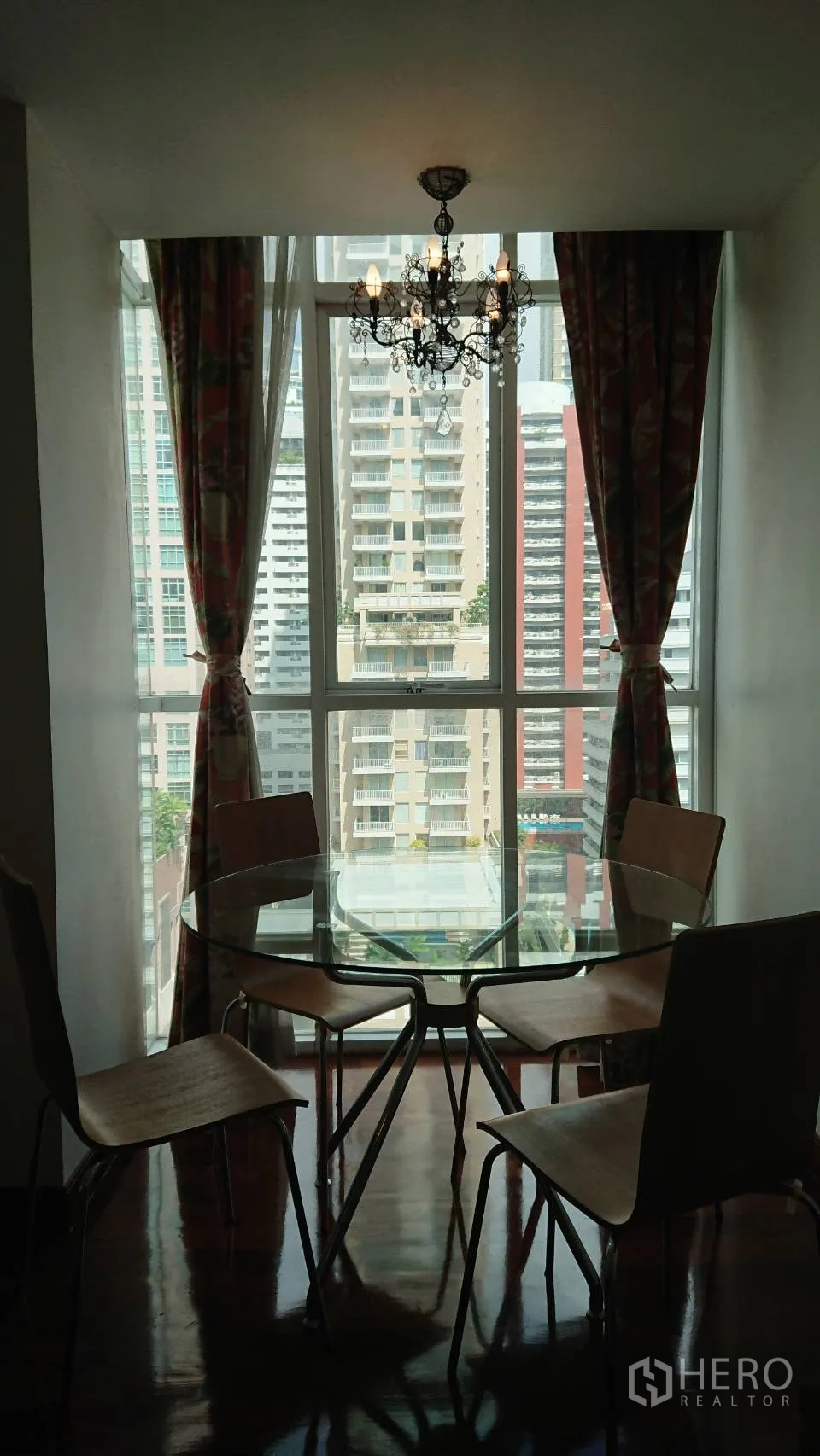 Condominium for rent in Pathum Wan, Bangkok - Dining nook with a round glass table and four chairs under a chandelier beside a large window overlooking high-rises.