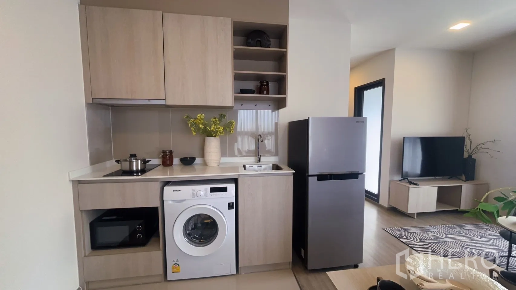 Condominium for rent in Watthana, Bangkok - Modern kitchenette with cooktop, sink, washing machine, microwave, and stainless refrigerator beside the living area.