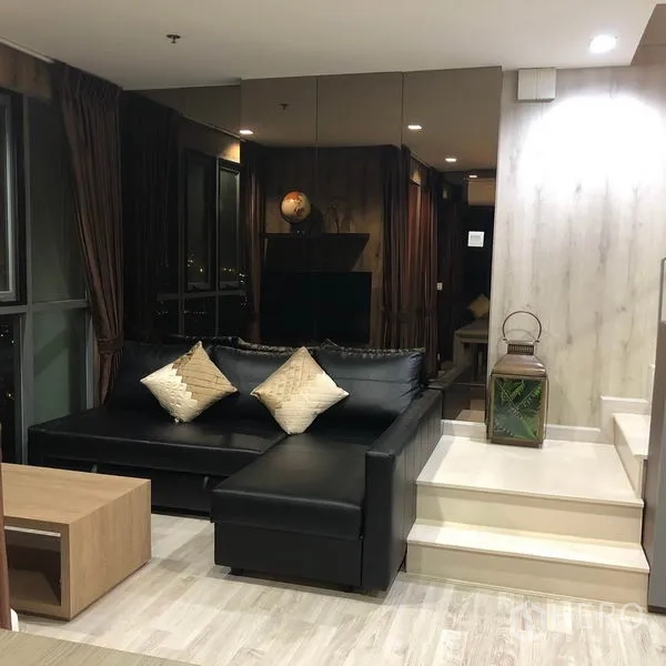 Condominium for rent or sale in Phra Khanong, Bangkok - Night view of the living room with a black L-shaped sofa, mirrored wall and raised platform.
