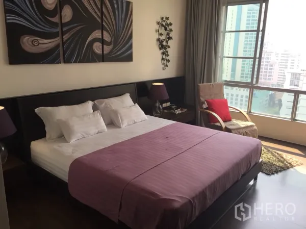 คอนโด ให้เช่าในKhlong Toei, Bangkok - ห้องนอนใหญ่เตียงคิงไซส์ โคมไฟข้างเตียง งานศิลป์ผนัง และหน้าต่างบานใหญ่เห็นวิวเมือง.