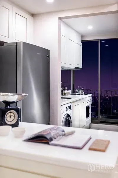 Condominium for rent in Huai Khwang, Bangkok - Bright kitchen with white cabinets, stainless fridge, washer and a balcony showing night city views.