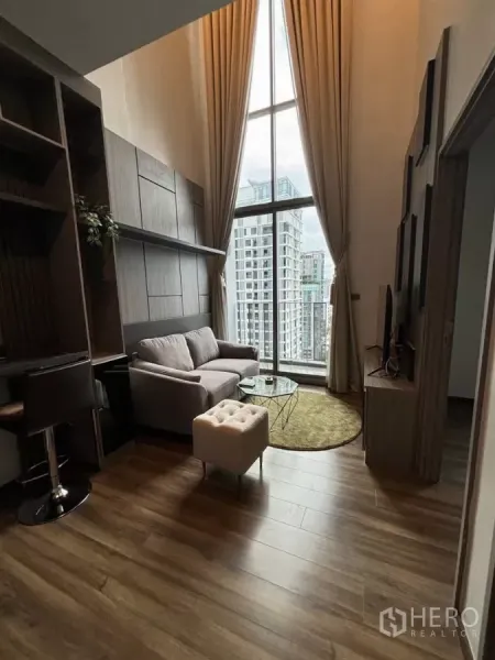 Condominium for rent or sale in Watthana, Bangkok - Cozy living area with sofa, round rug and tall windows offering city views.