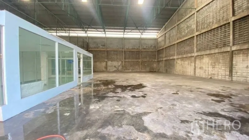 货栈 出租在Prawet, Bangkok - 仓库一角展示玻璃封闭办公室、抛光混凝土地面及配有照明的钢桁架屋顶。