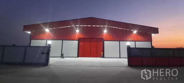 货栈 出租或出售在Khlong Luang, Pathum Thani - 傍晚正立面，宽大的红色推拉门、泛光灯与宽阔车道。