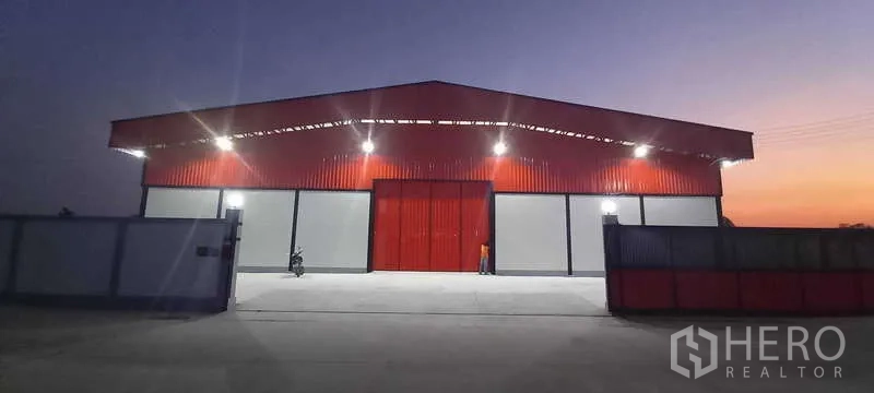 货栈 出租或出售在Khlong Luang, Pathum Thani - 傍晚正立面，宽大的红色推拉门、泛光灯与宽阔车道。