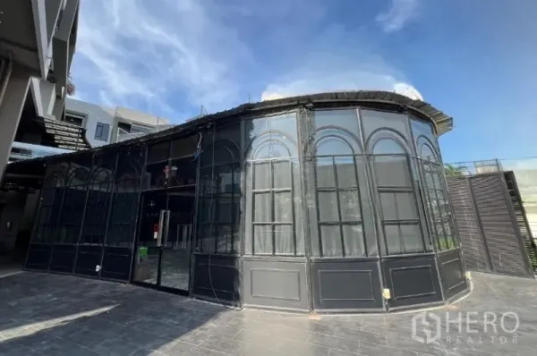 พื้นที่ขายปลีก ให้เช่า - หน้าร้านกระจกโค้งกรอบสีดำ พร้อมลานปูกระเบื้องด้านหน้า ใต้ท้องฟ้าสีฟ้า