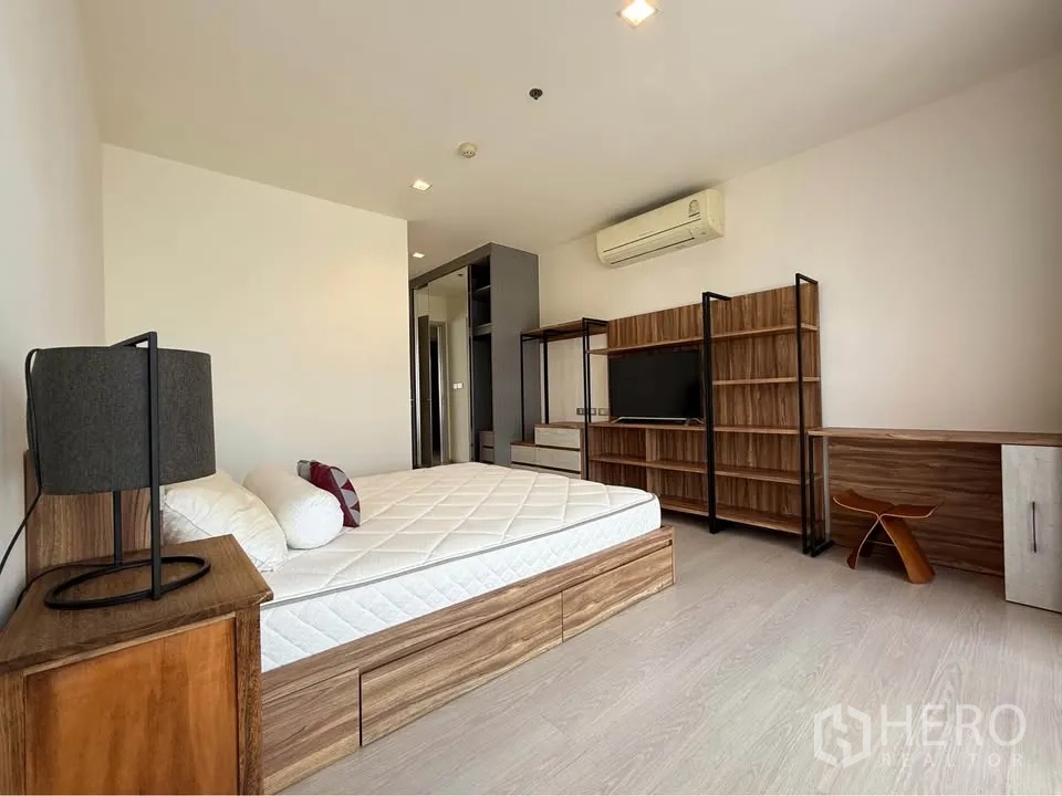 Condominium for rent in Khlong Toei, Bangkok - Sleeping area with bedframe, TV, shelving and desk by tall curtains.