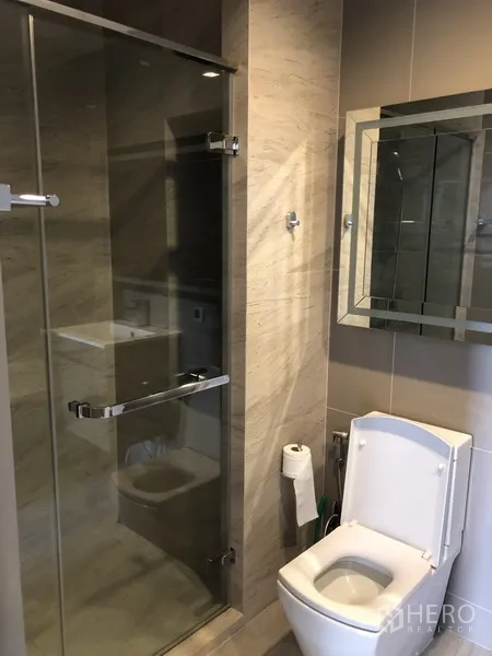 Condominium for rent in Watthana, Bangkok - Contemporary bathroom with a glass shower enclosure, mirror cabinet, and toilet in neutral tiles.
