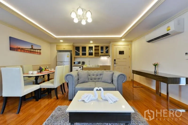 Apartment for rent in Khlong Toei, Bangkok - Open-plan living room with sofa, dining set and kitchenette under cove lighting.